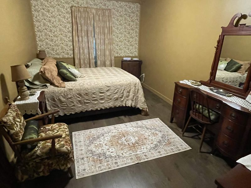 A bedroom with floral wallpaper behind the bed, made up with a cream patterned coverlet and green and tan pillows. A floral armchair and a nightstand with a lamp sit to the left, and a wooden vanity with a mirror and a chest of drawers stand to the right.
