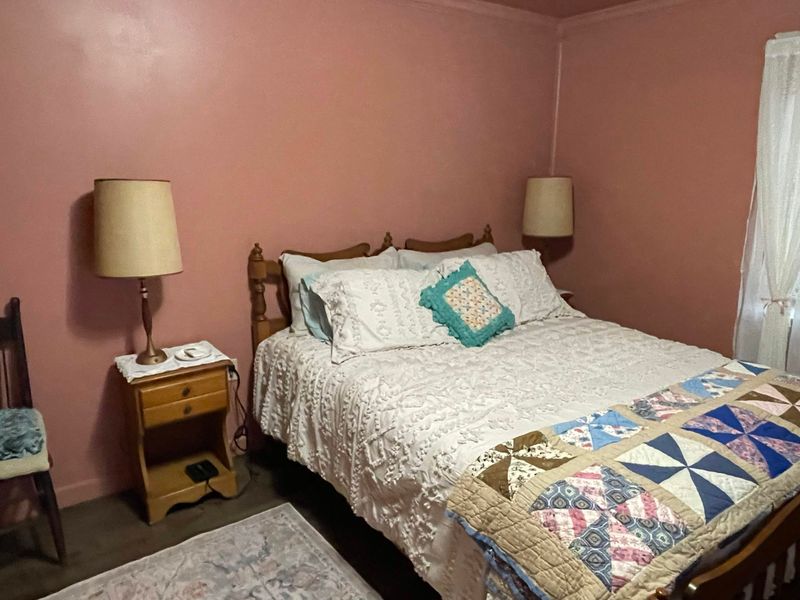 A bedroom with dusty pink walls and a wooden bed dressed in white bedding and a multicoloured geometric patchwork quilt, with a teal cushion on top. A nightstand and lamp stand to the left, and a window with white sheer curtains is on the right.