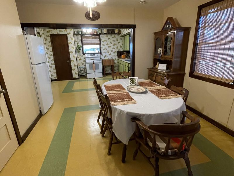 A dining table with a white cloth and woven placemats, ringed by dark wooden spindle-back chairs, one turned towards the viewer in the foreground, on a tan floor patterned with wide green bands. A white refrigerator and a door stand on the left, a tall glass-fronted wooden cabinet and a blinded window on the right, and an open pass-through behind the table reveals a kitchen with floral wallpaper.