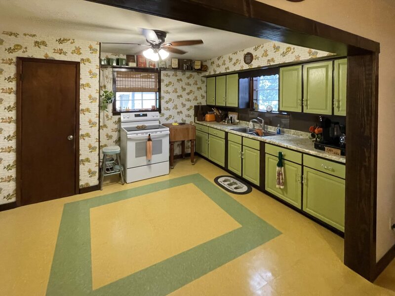 A kitchen framed by a dark wooden beam, with yellow flooring outlined by a green square and floral wallpaper. Light green cabinets with a sink under a window run along the right, a white stove stands beneath a second window, and a ceiling fan hangs overhead.