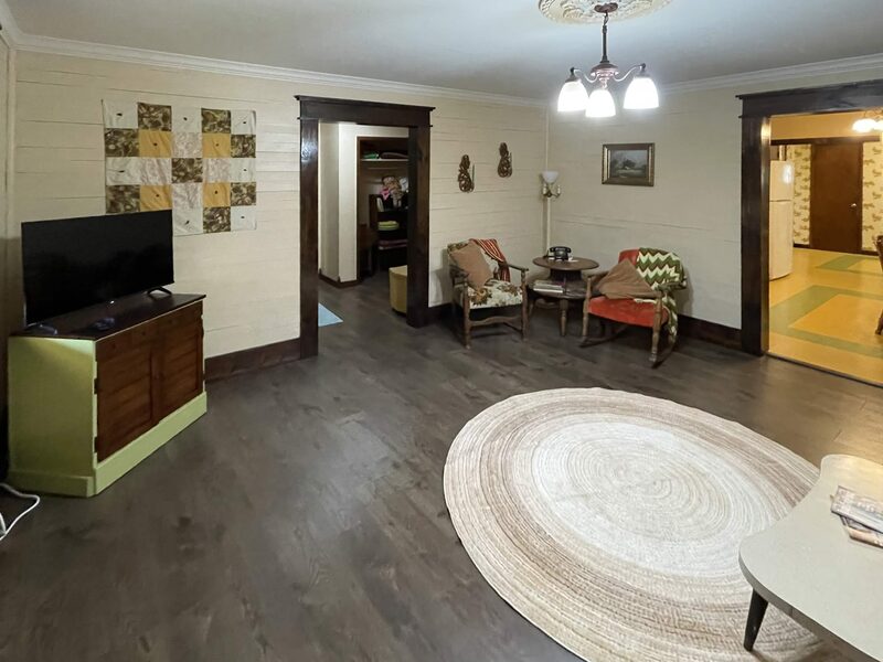 A living room where a flat-screen television stands on a low green and wood cabinet beneath a hanging patchwork quilt. A round woven rug with a small white table sits in the middle, and two dark-trimmed doorways lead to a hallway and to a kitchen with checkered tile.